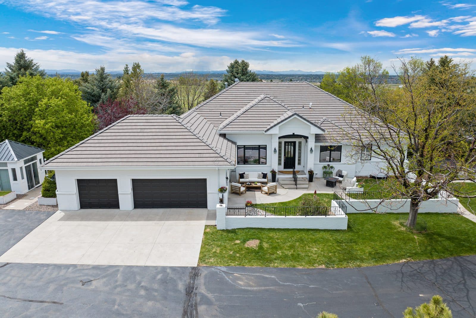 A Douglas County property from the air: a white single-story house with a tiled hip roof and a three-car garage, a wide concrete drive, low white garden walls around a lawn, mature trees on every side and the Front Range along the horizon.