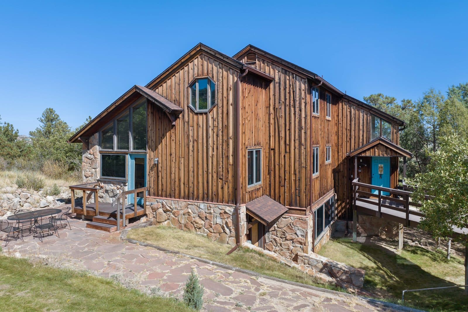 A Douglas County house clad in weathered vertical cedar over a rough stone base, with a steep gabled roof, turquoise-painted door and window frames, a flagstone patio with a small iron table, and dry native grass running out to ponderosa pines behind.