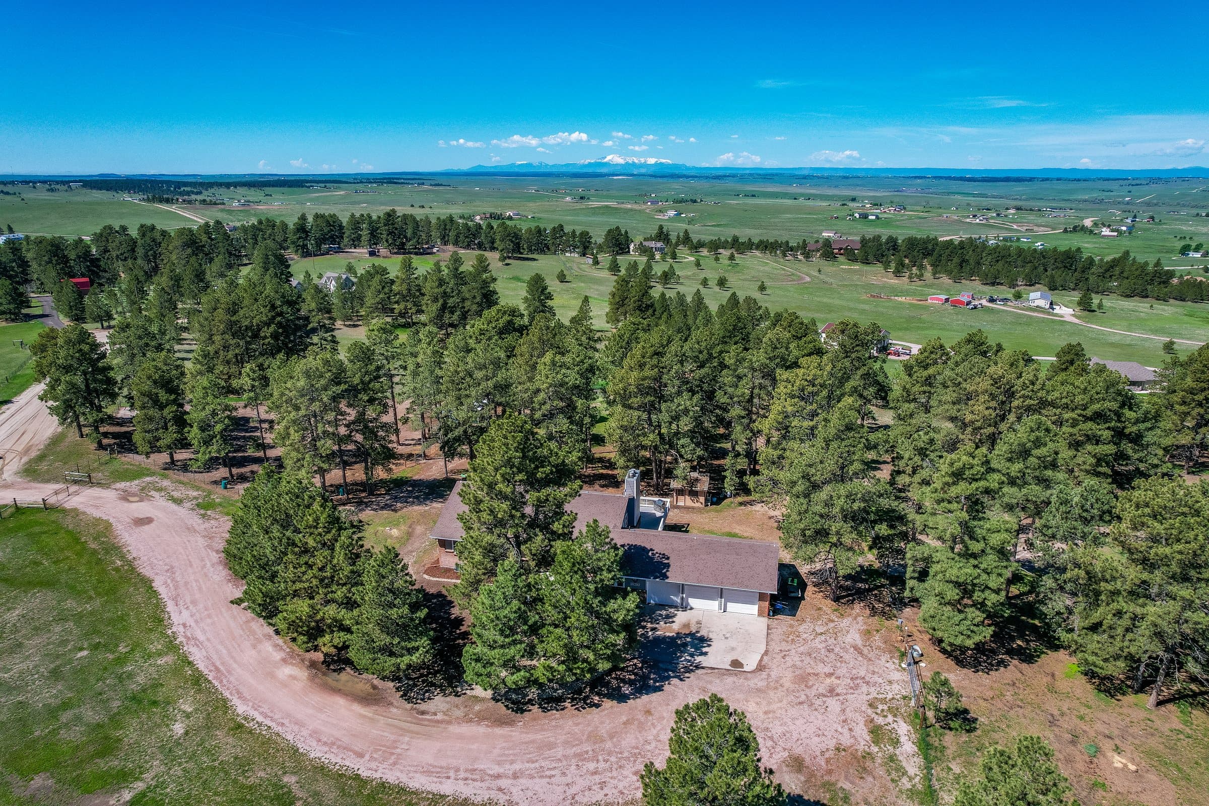 An Elbert County acreage lot seen from the air: a house among ponderosa pines, a curving dirt drive, fenced grassland running to the horizon and snow still on the peaks beyond.