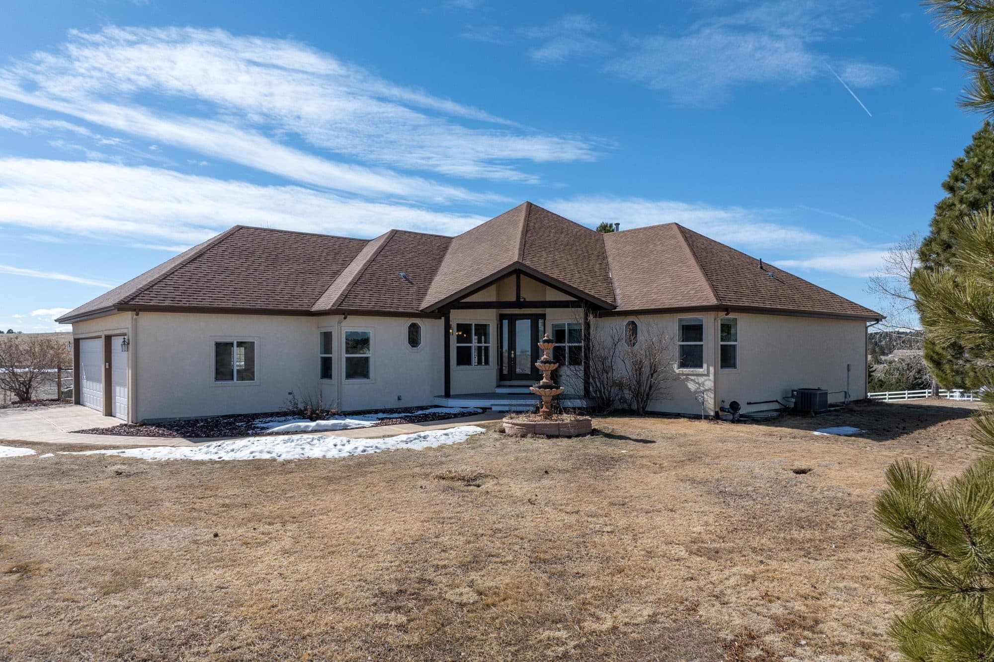 A single-story Elbert County ranch home with a stucco exterior and a hipped roof, a stone fountain in the front yard, and a split-rail fence and ponderosa pines at the edge of the lot.