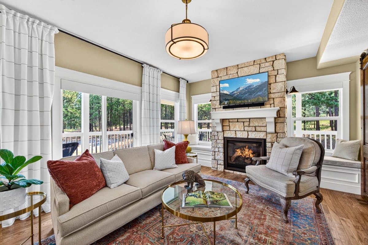 A living room with a floor-to-ceiling stone fireplace, a window seat, and tall windows looking out onto pines and a covered porch.