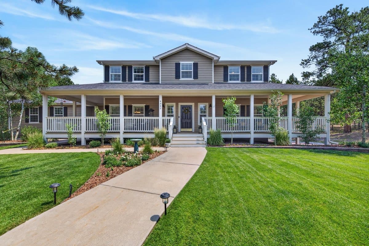 A two-story house seen from its front lawn, with a covered porch wrapping around both sides and pines behind it.